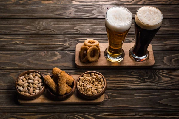 Copos de cerveja escura e leve com espuma perto de tigelas com amendoim, pistache, queijo frito e anéis de cebola na mesa de madeira — Fotografia de Stock