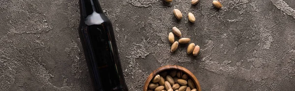 Панорамный снимок бутылки тёмного пива возле фисташек на коричневой текстурированной поверхности — стоковое фото