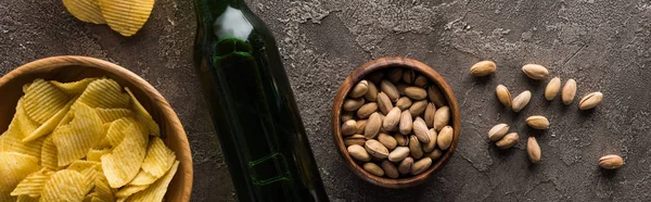 Colpo panoramico di bottiglia di birra vicino a ciotole con pistacchi e patatine su superficie martellata marrone — Foto stock