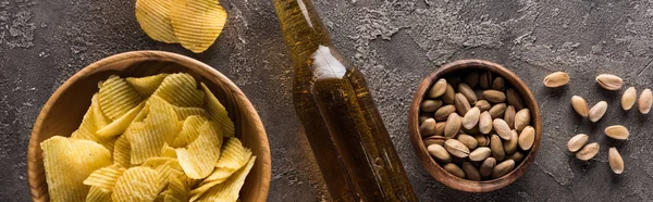 Panoramic shot of bottle of light beer near bowls with pistachios and crisps on brown textured surface — Stock Photo