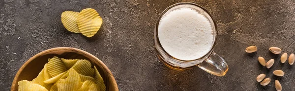 Foto panoramica di tazza di birra leggera vicino a patatine e pistacchi sparsi su una superficie martellata marrone — Foto stock