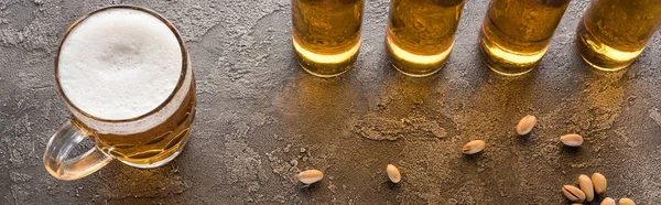 Panoramic shot of bottles of light beer near scattered pistachios on brown textured surface — Stock Photo