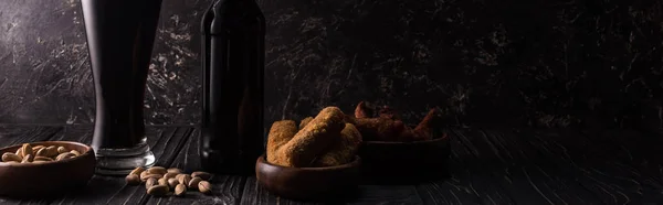 Panoramic shot of bottle and glass of dark beer near bowls with snacks on wooden table — Stock Photo