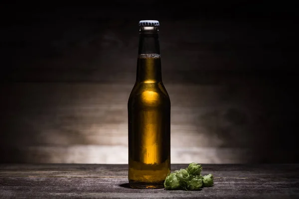 Bier in Flasche mit Hopfen auf Holztisch im Dunkeln bei Gegenlicht — Stockfoto