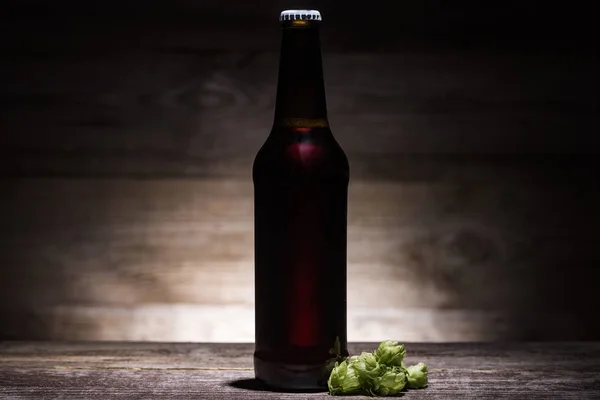 Bier in Flasche mit grünem Hopfen auf Holztisch im Dunkeln mit Gegenlicht — Stockfoto