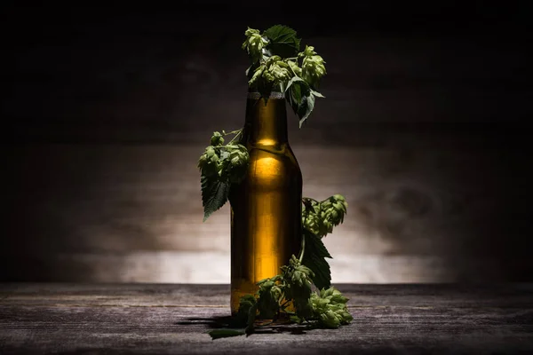 Bier in der Flasche mit frischem Hopfen auf Holztisch im Dunkeln bei Gegenlicht — Stockfoto