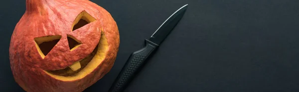Panoramaaufnahme eines Halloween-Kürbisses in der Nähe eines Messers auf schwarzem Hintergrund — Stockfoto