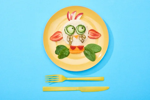 Top view of plate with fancy face made of food for childrens breakfast near cutlery on blue background — Stock Photo