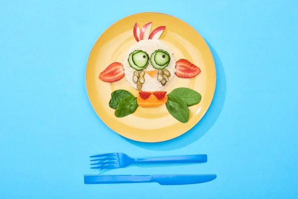 Top view of plate with fancy face made of food for childrens breakfast near cutlery on blue background — Stock Photo