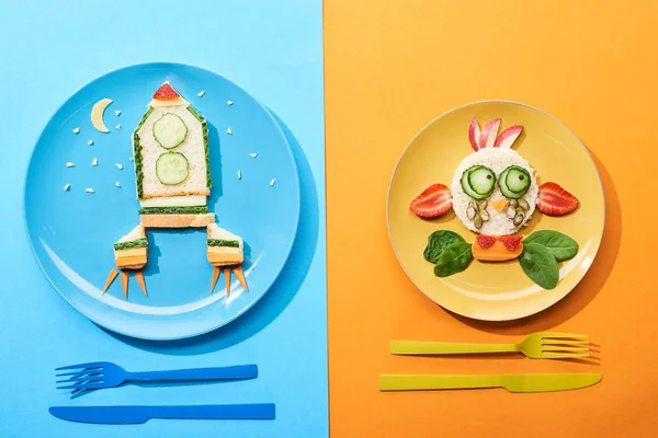 Top view of plates with fancy face and rocket made of food for childrens breakfast near cutlery on blue and orange background — Stock Photo