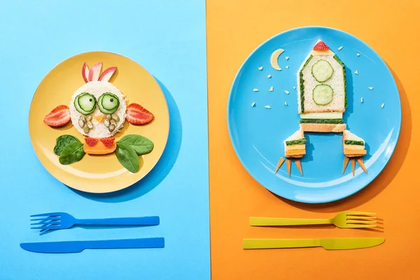 Top view of plates with fancy face and rocket made of food for childrens breakfast near cutlery on blue and orange background — Stock Photo