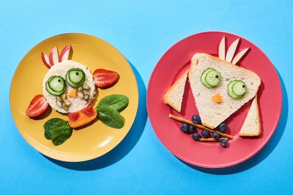 Vista superior de platos con vaca y pájaro de lujo hechos de comida para niños desayuno sobre fondo azul - foto de stock