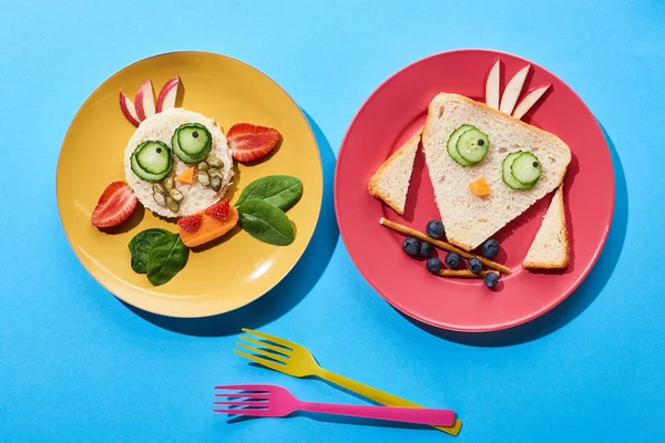 Vue de dessus des assiettes avec vache de fantaisie et oiseau fait de nourriture pour le petit déjeuner des enfants près des fourches sur fond bleu — Photo de stock