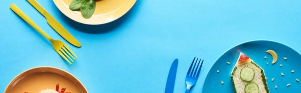 Top view of plates with fancy childrens breakfast near cutlery on blue background, panoramic shot — Stock Photo