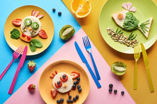 Vista dall'alto di piatti con animali di fantasia fatti di cibo per bambini colazione su sfondo blu, giallo e rosa con forchette e coltelli — Foto stock