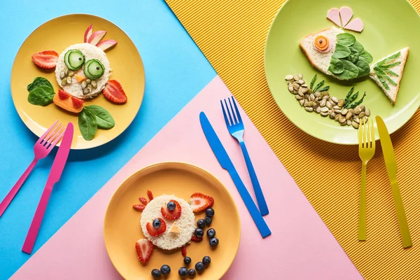 Top view of plates with fancy animals made of food for childrens breakfast on blue, yellow and pink background with cutlery — Stock Photo