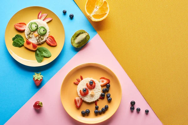 Vista superior de platos con animales de lujo hechos de comida para niños desayuno sobre fondo azul, amarillo y rosa - foto de stock