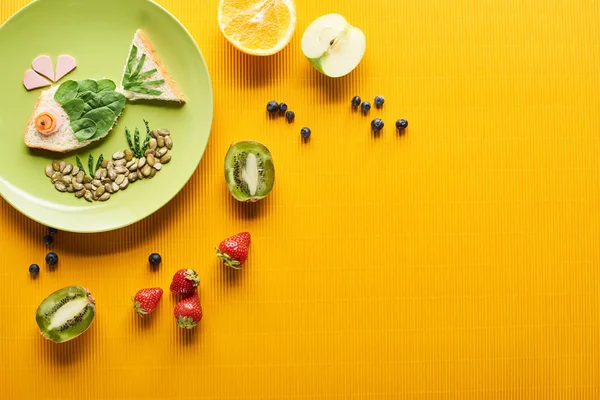 Top view of plate with fancy fish made of food near scattered fruits on colorful orange background — Stock Photo