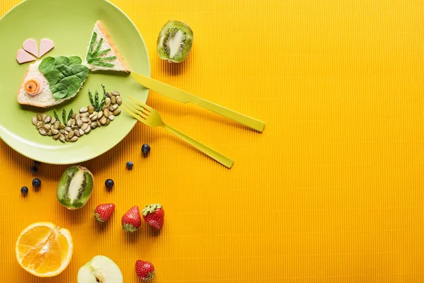 Вид сверху на тарелку с модной рыбой, сделанной из еды рядом с фруктами и столовыми приборами на красочном оранжевом фоне — стоковое фото