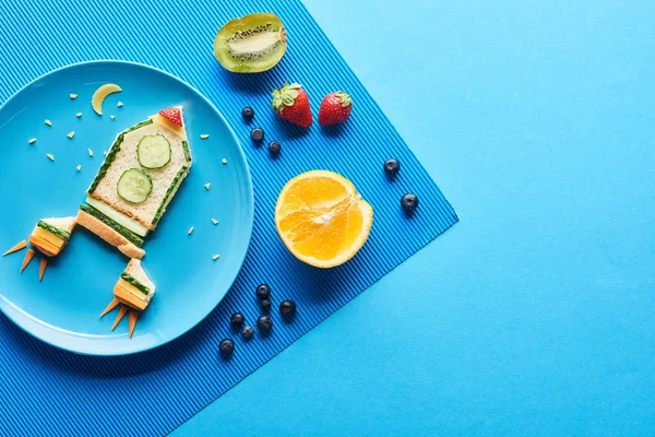 Vista dall'alto di piatti con razzo fantasia fatta di cibo su sfondo blu con frutta — Foto stock
