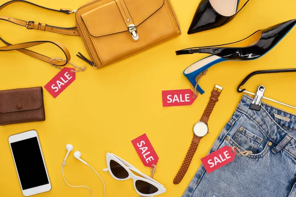 Вид на модную одежду и аксессуары с этикетками и смартфоном с наушниками на желтом фоне — стоковое фото