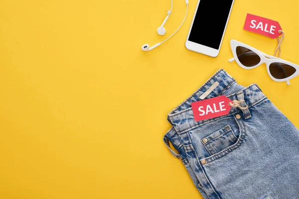 Vista superior de jeans, gafas y smartphone con auriculares con etiquetas de venta sobre fondo amarillo - foto de stock