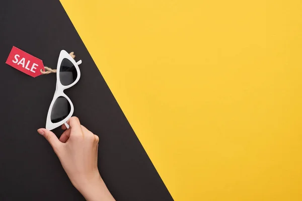 Vista recortada de mujer sosteniendo gafas con etiqueta de venta sobre fondo amarillo y negro - foto de stock