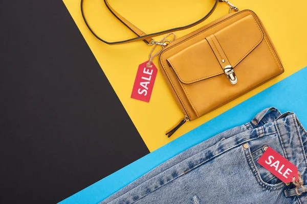 Top view of bag and jeans with sale labels on blue, yellow and black background — Stock Photo