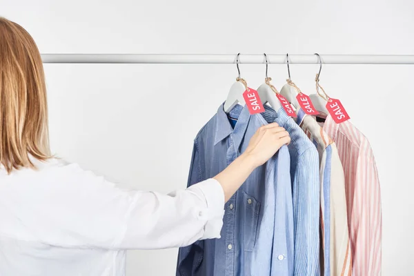 Vista ritagliata di donna bionda vicino a camicie eleganti appese con etichette di vendita isolate su bianco — Foto stock