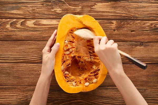 Cropped view of woman taking out seeds from half of pumpkin on brown wooden surface — Stock Photo