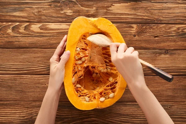 Vista recortada de la mujer sacando semillas de la mitad de la calabaza en la superficie de madera marrón - foto de stock