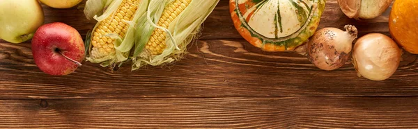 Panoramic shot of raw sweet corn, pumpkins, onions and apples on brown wooden surface — Stock Photo