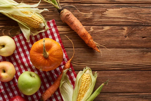 Vista dall'alto della tovaglia a quadretti con zucca, mais dolce, mele e carote su superficie di legno con spazio per copiare — Foto stock