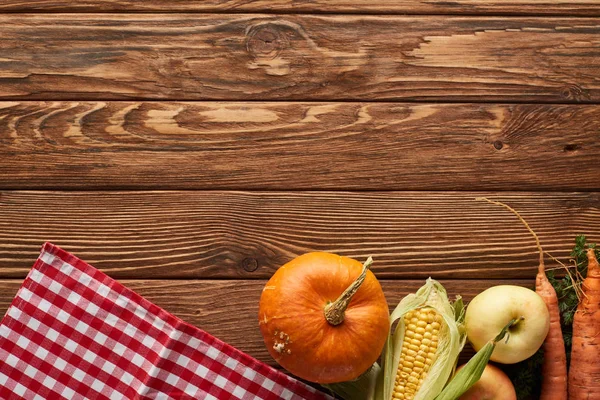 Top view of checkered tablecloth near pumpkin, sweet corn, apples and carrots on wooden surface with copy space — Stock Photo