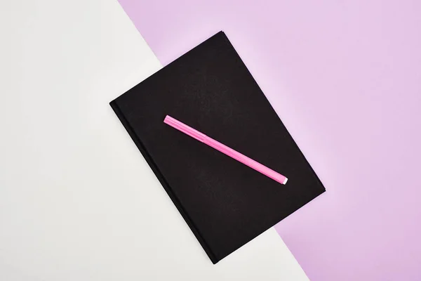 Tendido plano con cuaderno negro con pluma de fieltro rosa sobre fondo violeta y blanco - foto de stock