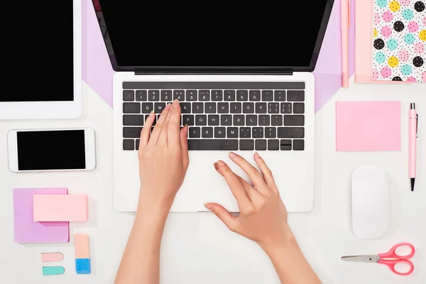 Vista parcial de la mujer usando el ordenador portátil cerca de gadgets y suministros de oficina sobre fondo violeta y blanco - foto de stock