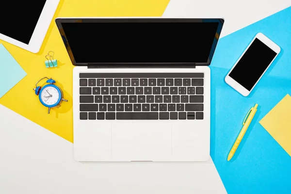 Вид зверху на ноутбук, смартфон, цифровий планшет з порожнім екраном з офісними приладдям на жовтому, синьому та білому тлі — стокове фото