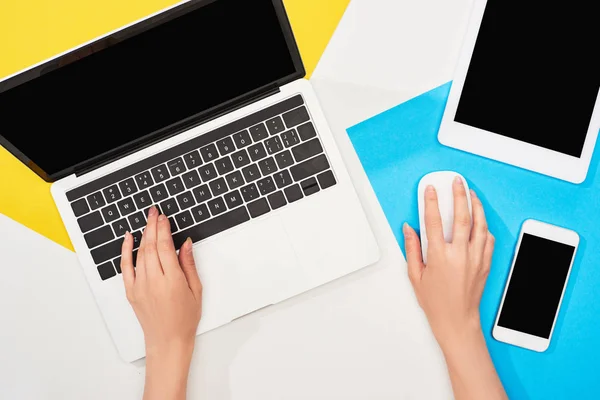 Cropped view of woman using laptop and computer mouse near smartphone, digital tablet on yellow, blue and white background — Stock Photo