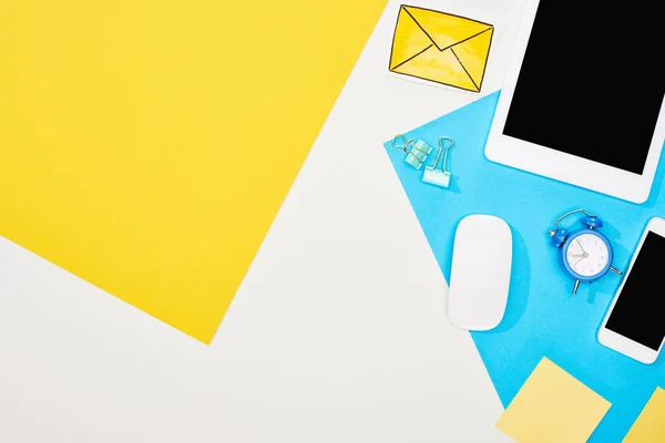 Вид сверху смартфона, цифровой планшет с пустым экраном и компьютерная мышь с офисными принадлежностями на желтом, синем и белом фоне — стоковое фото