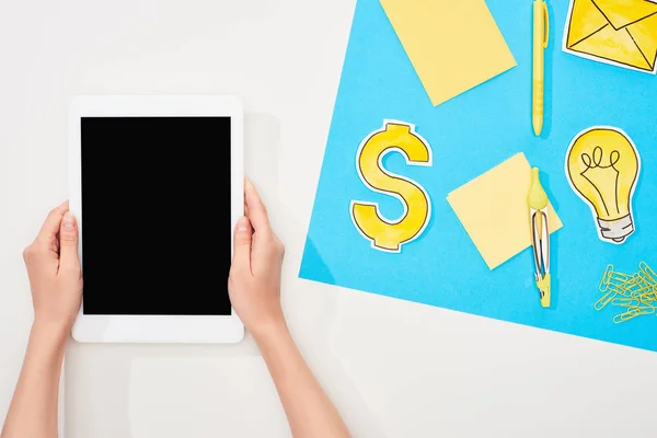 Cropped view of woman holding digital tablet at workplace with office supplies, yellow paper icons on blue and white background — Stock Photo