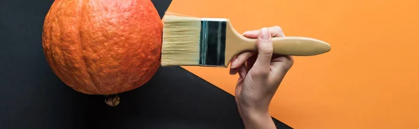 Panoramic shot of woman holding paintbrush near pumpkin on black and orange background — Stock Photo