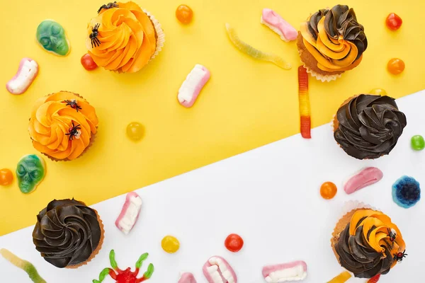Vista superior de coloridos dulces y cupcakes gomosos sobre fondo amarillo y blanco con espacio para copiar, regalo de Halloween - foto de stock