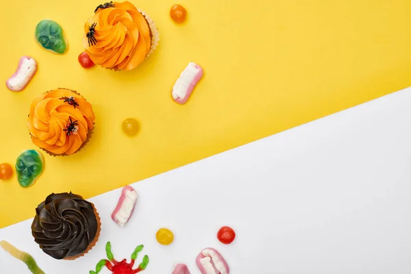 Vue de dessus de bonbons gommeux colorés et cupcakes sur fond jaune et blanc, festin d'Halloween — Photo de stock