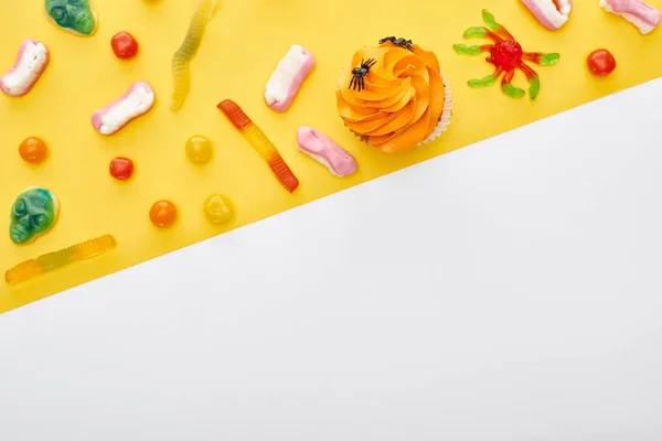 Vista superior de coloridos dulces gomosos y cupcake sobre fondo amarillo y blanco con espacio para copiar, regalo de Halloween - foto de stock