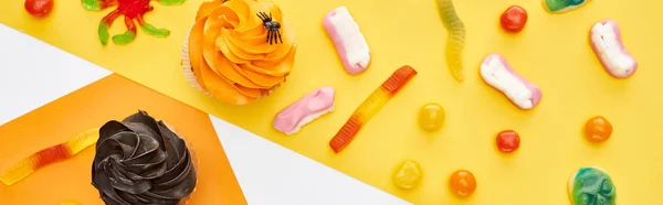 Draufsicht auf bunte Gummibärchen und Cupcakes auf gelb-weißem Hintergrund, Halloween-Leckerbissen — Stockfoto