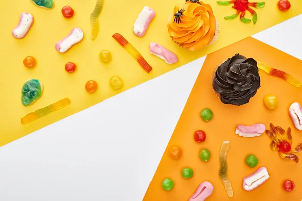 Vue de dessus de bonbons gommeux colorés et cupcakes sur fond jaune et blanc, festin d'Halloween — Photo de stock