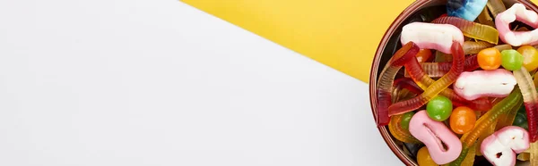 Plano panorámico de dulces gomosos coloridos en un tazón sobre fondo amarillo y blanco, regalo de Halloween - foto de stock