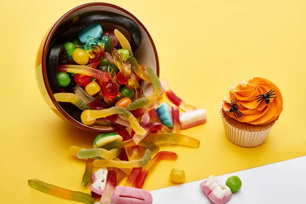 Colorful gummy sweets scattered from bowl near cupcake on yellow background, Halloween treat — Stock Photo