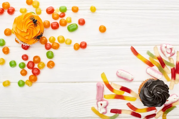 Draufsicht auf bunte Gummibonbons, Cupcakes und Bonbons auf weißem Holztisch, Halloween-Leckerbissen — Stockfoto