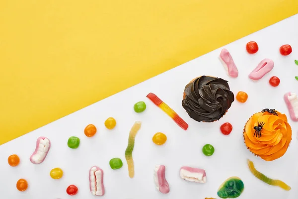 Draufsicht auf bunte Gummiwürmer in der Nähe von Cupcakes auf weißem und gelbem Hintergrund, Halloween-Leckerbissen — Stockfoto